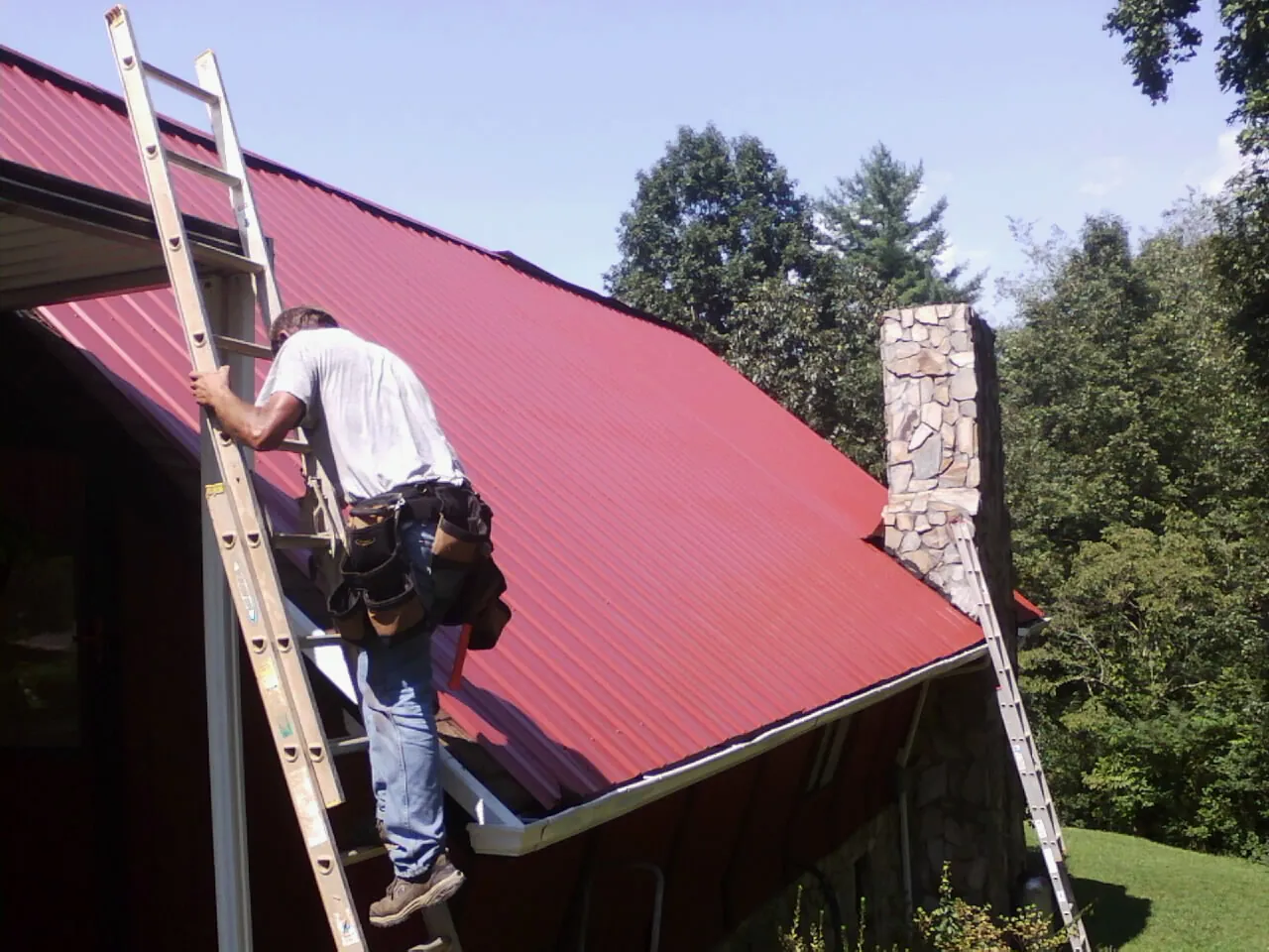 Roofer on a ladder beside a red metal roof and stone chimney — Soffit & Fascia Repair by Roofing Brothers in Bakersville